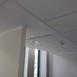 Multifinish - Pomieszczenie biurowe po odświeżeniu: świeżo pomalowane ściany w odcieniach bieli, sufit kasetonowy z zamontowanymi lampami i czujnikiem dymu, widok z narożnika, naturalne światło wpadające przez...