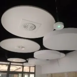 Białe, okrągłe panele akustyczne zamontowane na czarnym suficie podwieszanym, widoczne kratki wentylacyjne i oświetlenie punktowe. Jeden z paneli ma kwadratową kratkę wentylacyjną pośrodku.