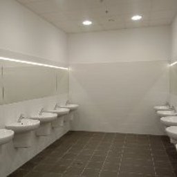 Multifinish - Wnętrze publicznej toalety z rzędem białych umywalek w ceramicznych misach, podłużne lustra z oświetleniem LED, szara podłoga z płytek, minimalistyczny styl.