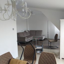 Multifinish - Wykończone wnętrze pokoju dziennego z widocznym narożnikiem, szklanym stolikiem, krzesłami z brązową tapicerką i metalowymi nogami, oraz dekoracyjnym żyrandolem w stylu retro.