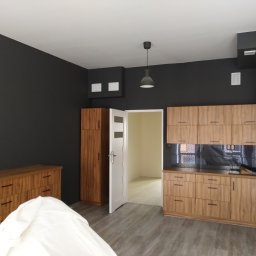 Multifinish - Wykończone wnętrze pokoju z drewnianymi meblami kuchennymi, czarną ścianą i szarą podłogą, widok na sąsiednie pomieszczenie przez otwarte drzwi.