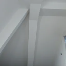 Pomalowane na biało poddasze ze skośnym sufitem i widocznymi belkami konstrukcyjnymi, fragment srebrnej lampy w prawym górnym rogu, okno dachowe w tle.