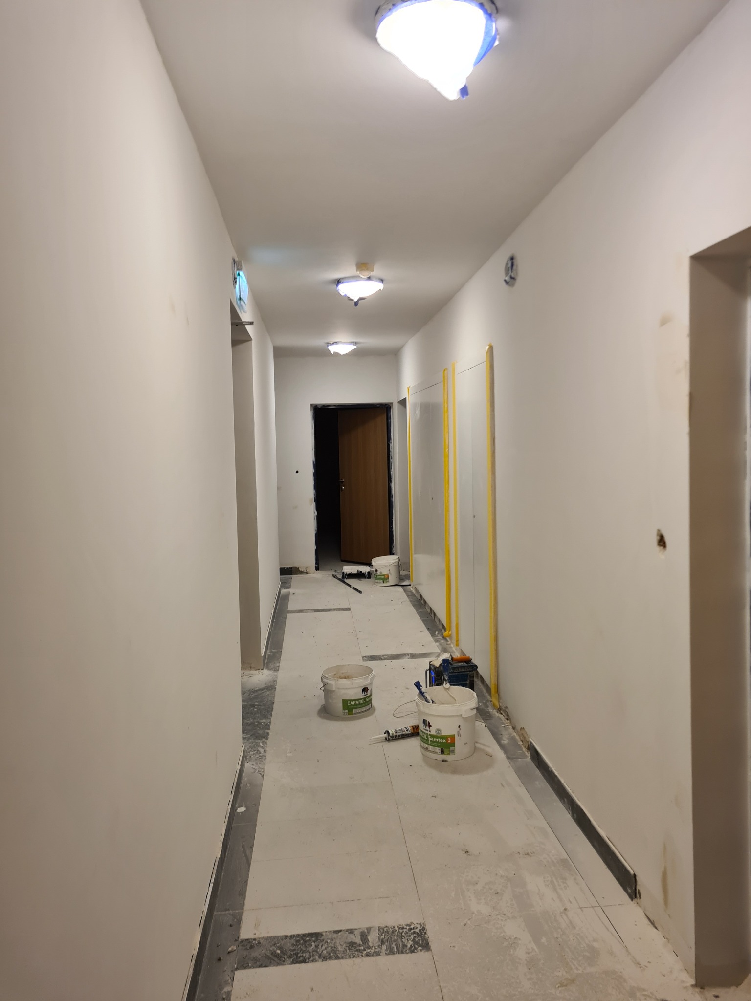 Korytarz w trakcie odświeżania ścian, widoczne białe ściany po nałożeniu gładzi, zabezpieczone drzwi, pędzle, wiaderka z farbą Caparol i Bamtex 3 na podłodze oraz taśma malarska na suficie.
