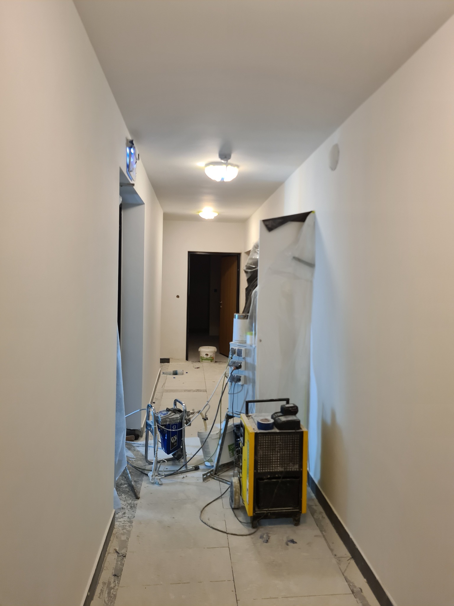 Korytarz w trakcie prac wykończeniowych: świeżo pomalowane, jasne ściany, widoczny agregat malarski, osuszacz powietrza, wiadro z farbą oraz zabezpieczone folią drzwi.