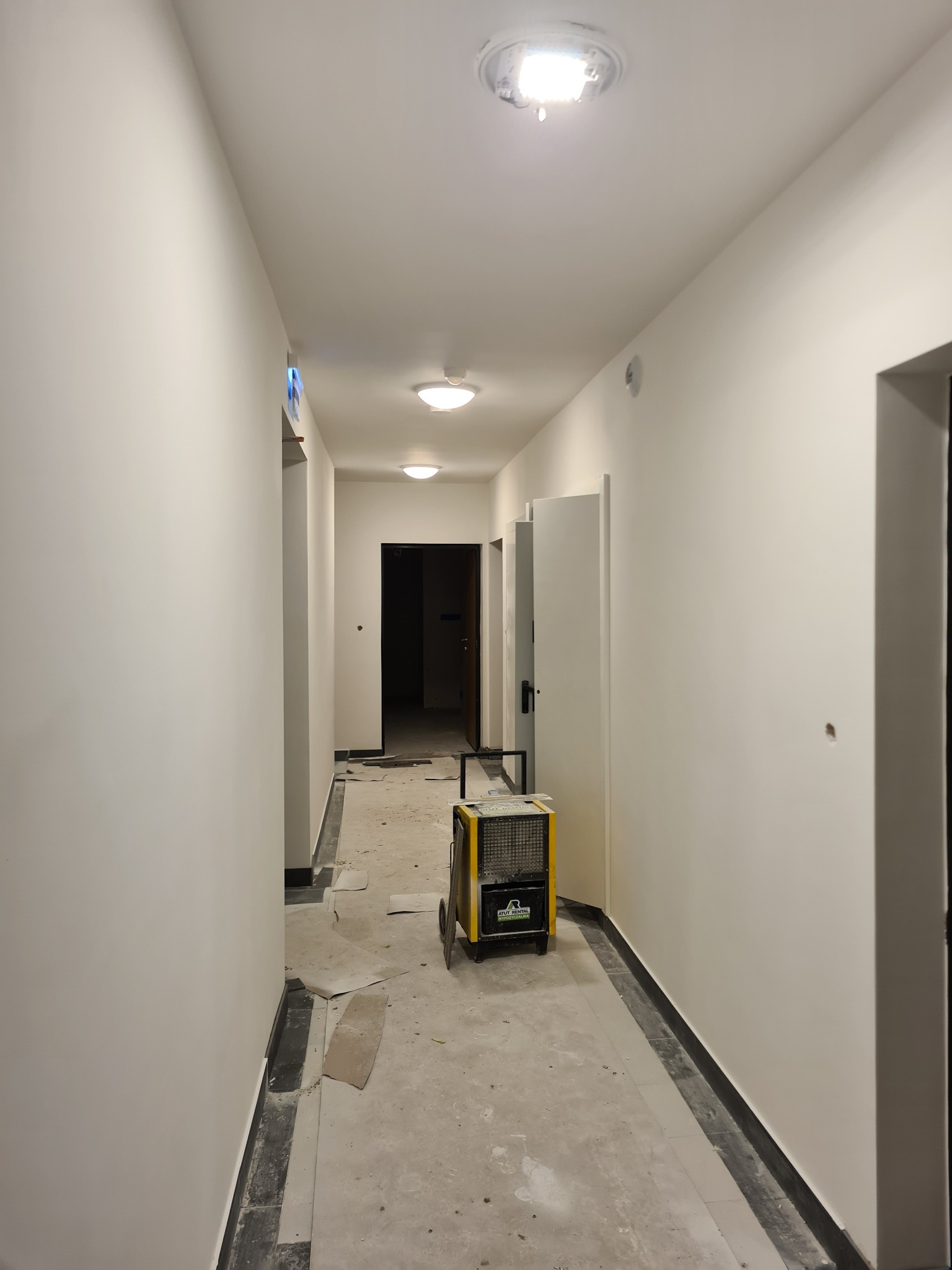 Korytarz w trakcie prac wykończeniowych; świeżo pomalowane, gładkie ściany, na podłodze resztki materiałów budowlanych, widoczny osuszacz powietrza.
