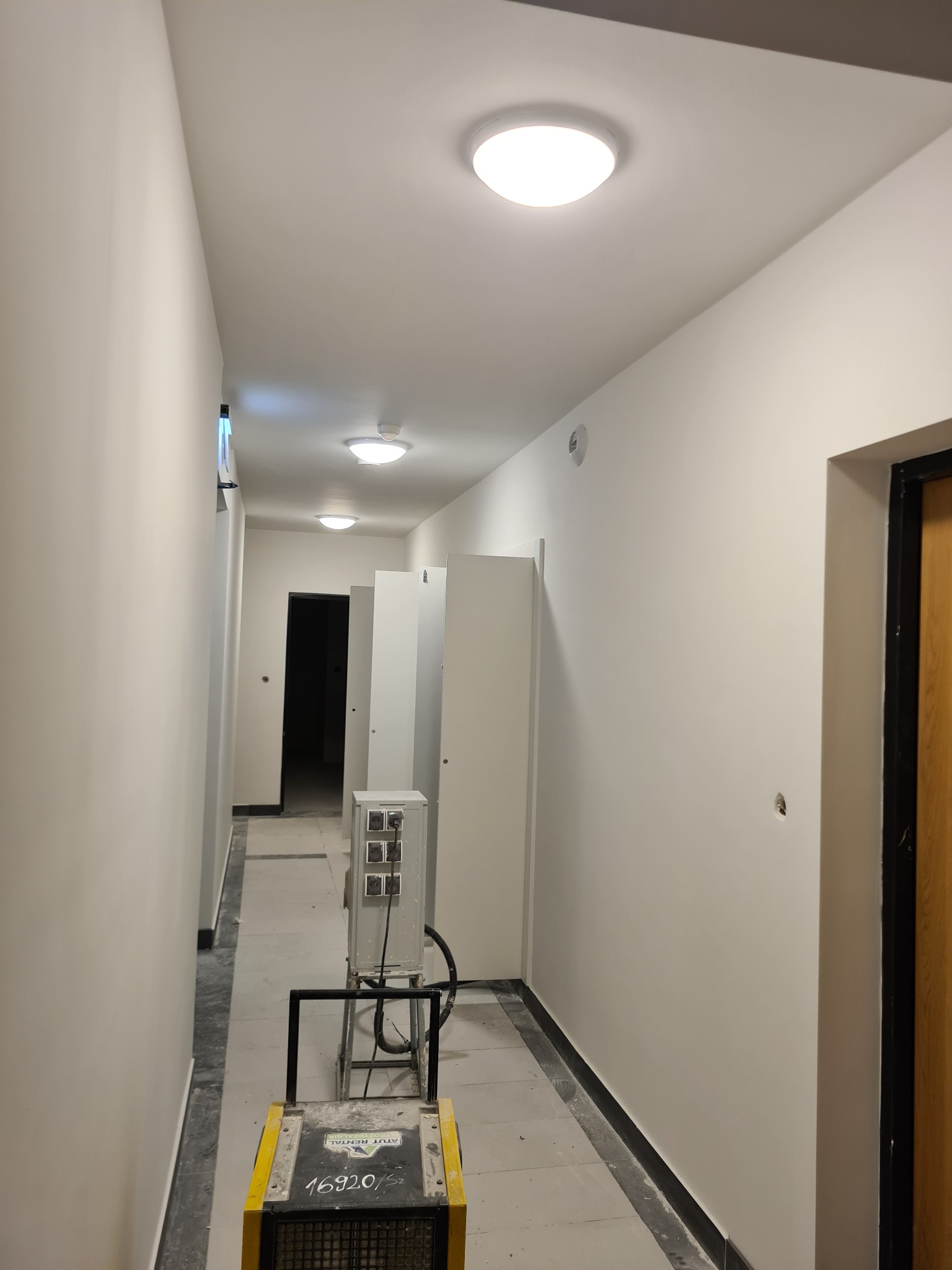 Korytarz z świeżo pomalowanymi ścianami na biało, widoczne trzy lampy sufitowe, wentylator przemysłowy na podłodze, przygotowany do dalszych prac wykończeniowych.