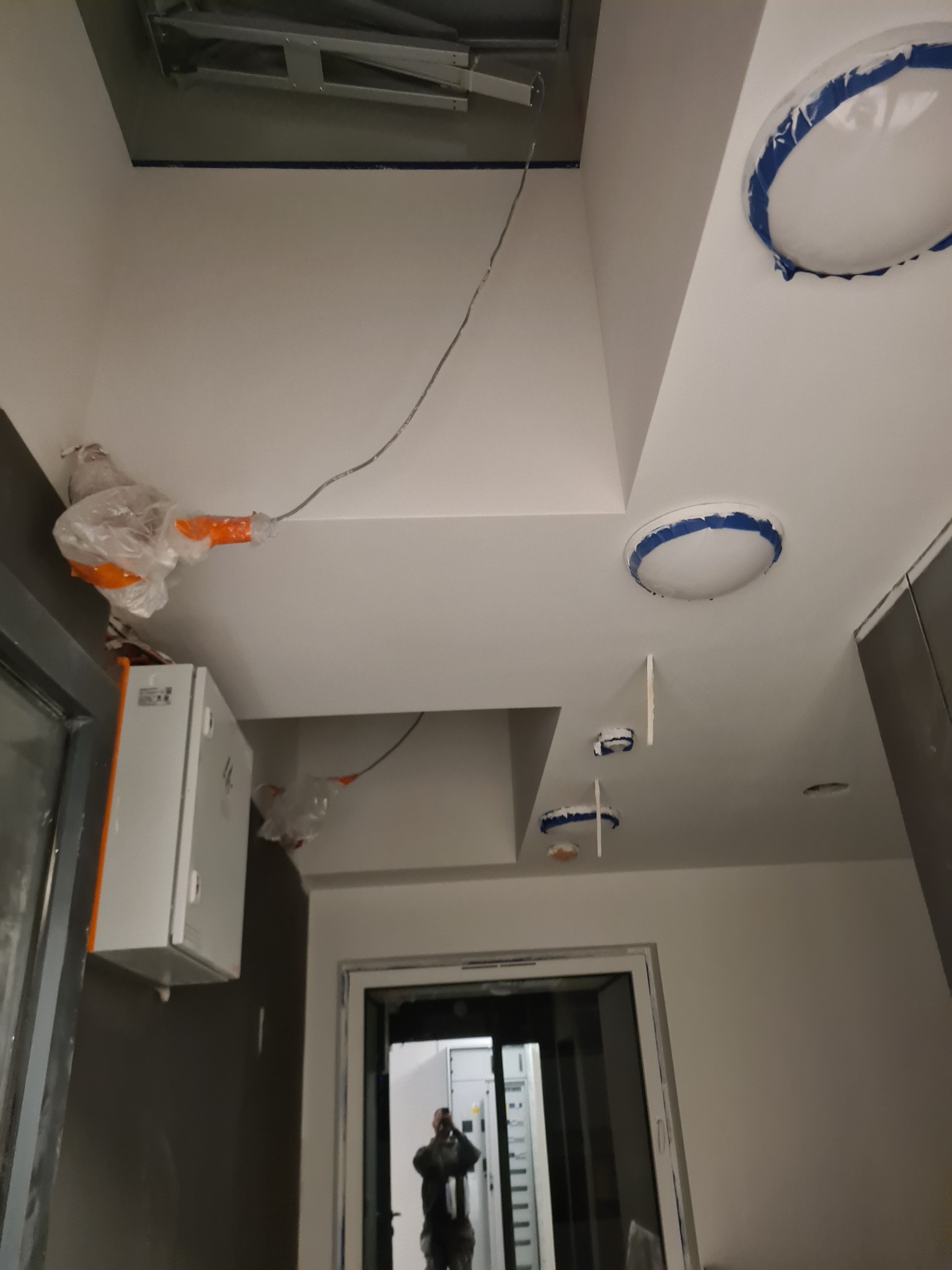 Przygotowanie do malowania sufitu: zabezpieczone taśmą malarską oprawy oświetleniowe, zakryte folią elementy, widoczne przewody i drabina rozłożona przy suficie.