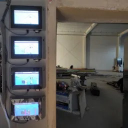 Cztery ekrany dotykowe zamontowane na ścianie, pokazujące schematy instalacji w budynku w stanie surowym. Widoczne przewody i puszki instalacyjne. W tle otwarty, surowy stan budynku z materiałami...