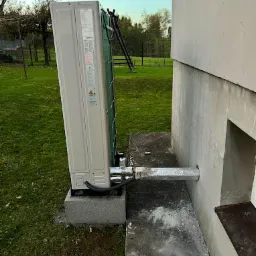 Zewnętrzna jednostka klimatyzacji zamontowana na betonowym fundamencie przy ścianie budynku, z widocznymi rurami i przewodami, otoczona trawnikiem i elementami krajobrazu.