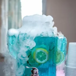Szklany dyspenser z niebieskim napojem, plasterkami cytryny i efektem suchego lodu, obok stos plastikowych kubków.
