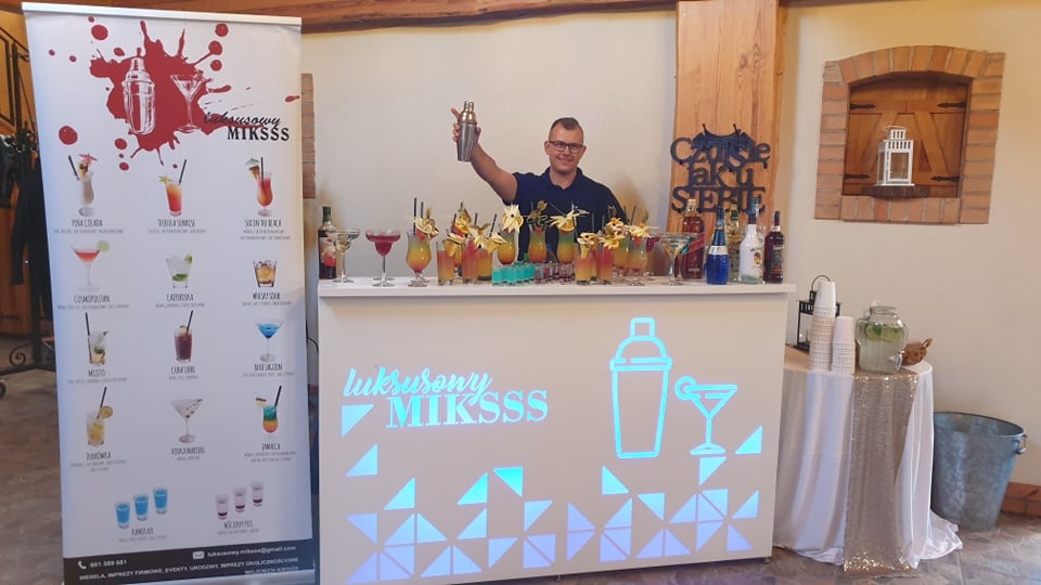 Barman za podświetlonym barem z drinkami, obok baner z menu drinków i dekoracje.