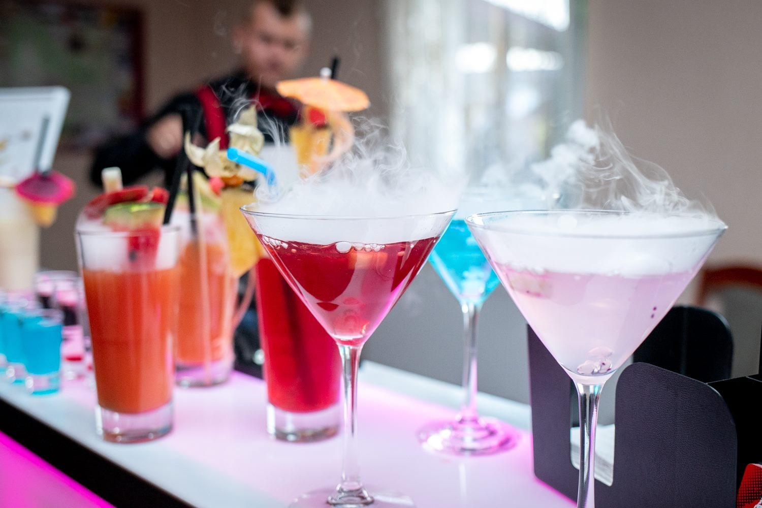 Bar z kolorowymi drinkami, z efektem suchego lodu, przygotowywany przez barmana w tle, oświetlony różowym światłem.