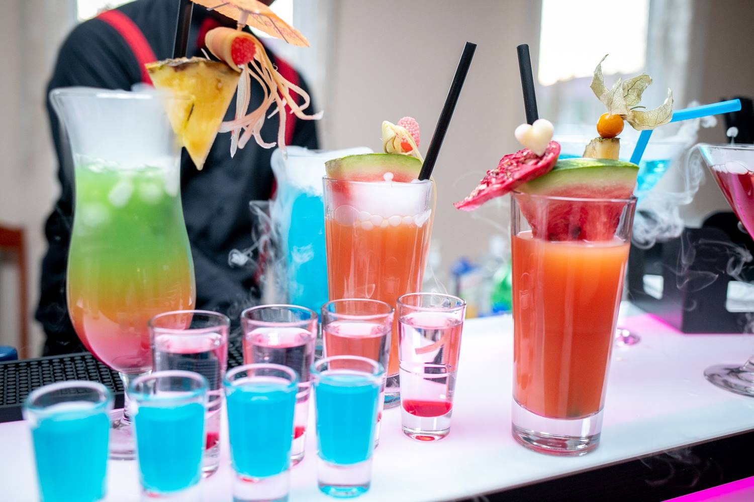 Barman w czarnej koszuli z szelkami przygotowuje kolorowe drinki z owocowymi dekoracjami i suchym lodem, na podświetlonym blacie.