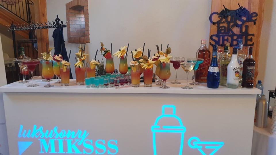 Bar z podświetlanym logo i drinkami z owocowymi dekoracjami, butelki alkoholu w tle, rustykalny wystrój wnętrza.