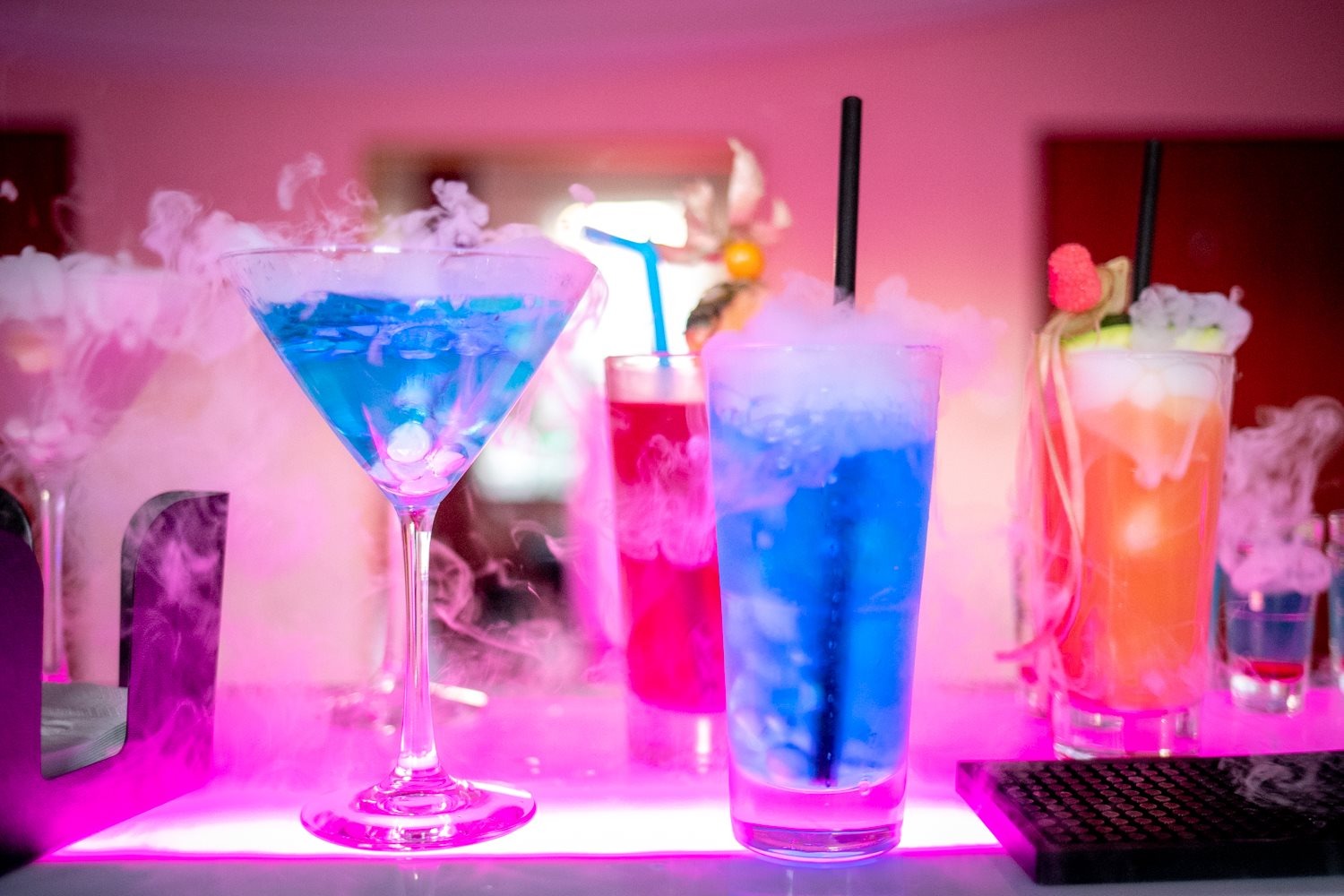 Bar z drinkami w różnych kolorach, otoczone suchym lodem, podświetlone różowym światłem