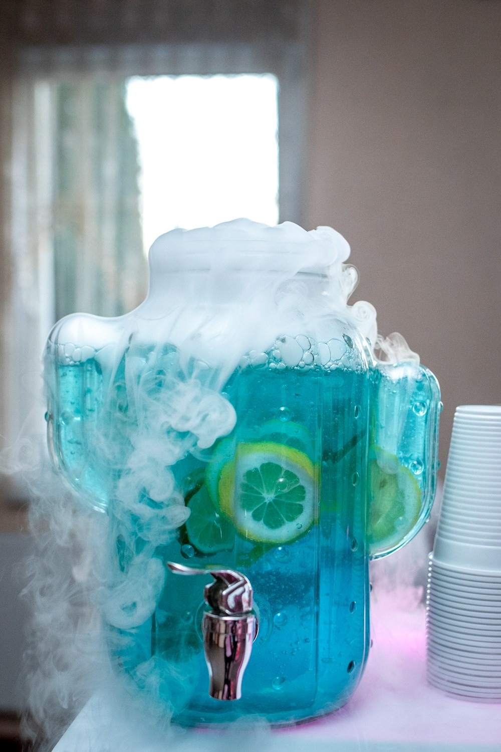 Szklany dyspenser z niebieskim napojem, plasterkami cytryny i efektem suchego lodu, obok stos plastikowych kubków.