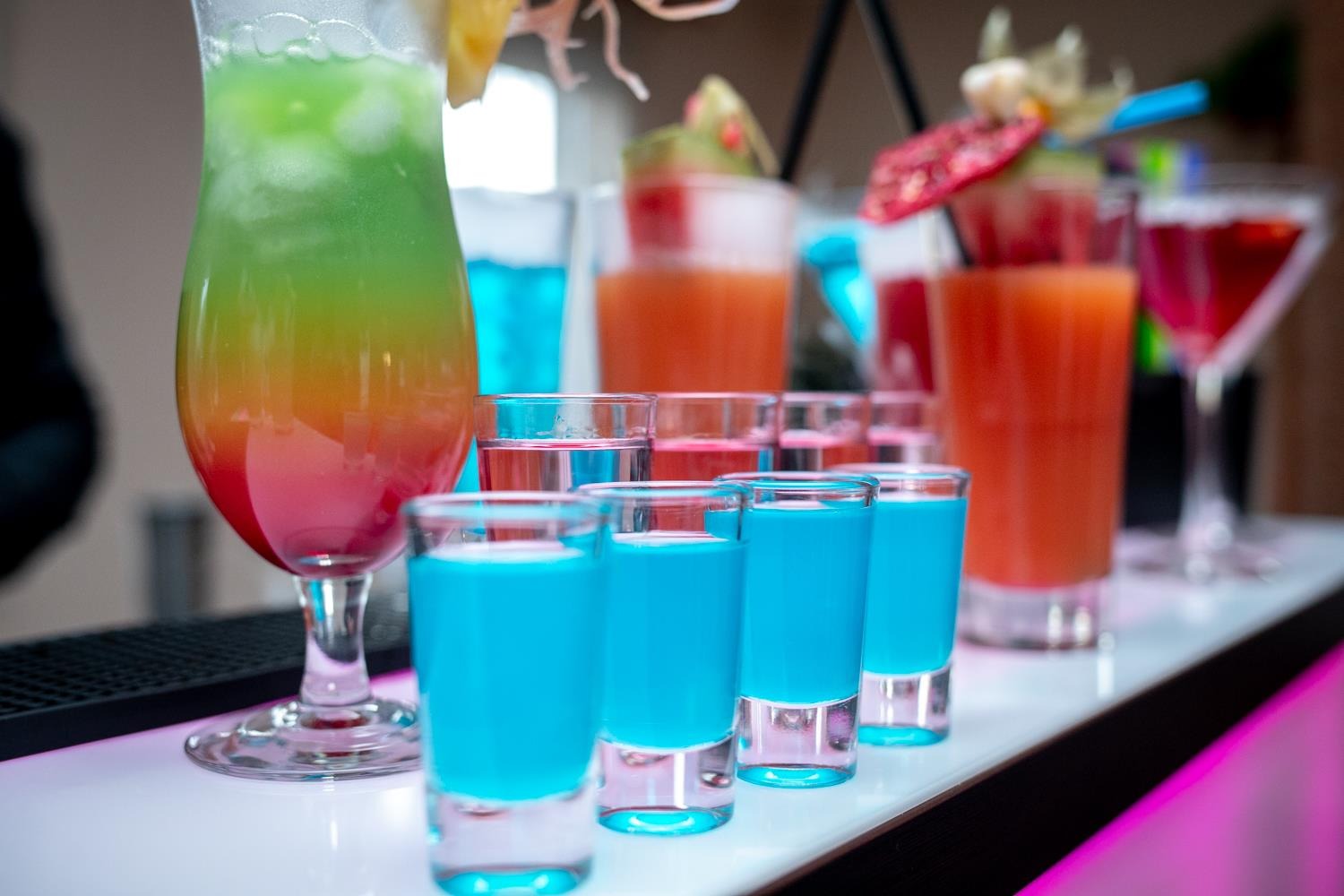 Rząd kolorowych drinków w szklankach o różnej wielkości, w tym wysoki koktajl z ananasem, na białym barze podświetlonym na różowo.