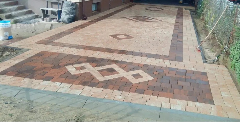 Ułożona kostka brukowa w dwóch odcieniach beżu i brązu, tworząca geometryczny wzór na podjeździe, z widocznym workiem z zaprawą w tle.