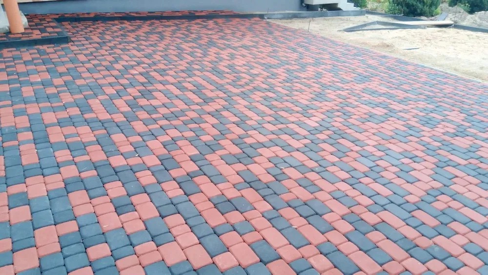 Ułożona kostka brukowa w kolorze czerwonym i grafitowym, tworząca wzór na podjeździe. Widoczny fragment budynku w tle.