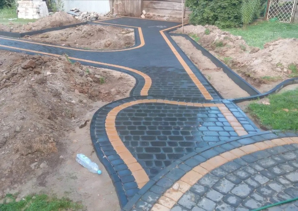 Ścieżka z kostki brukowej w odcieniach szarości i beżu, z widocznymi fragmentami terenu przygotowanego pod dalsze prace, butelka wody mineralnej na ziemi.