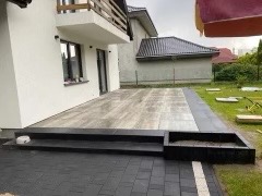 Wykończony taras z jasnoszarej kostki brukowej, obramowany ciemnoszarą kostką, przylegający do białego budynku z drewnianą balustradą. Widoczne schody prowadzące na taras oraz donica z ciemnej...