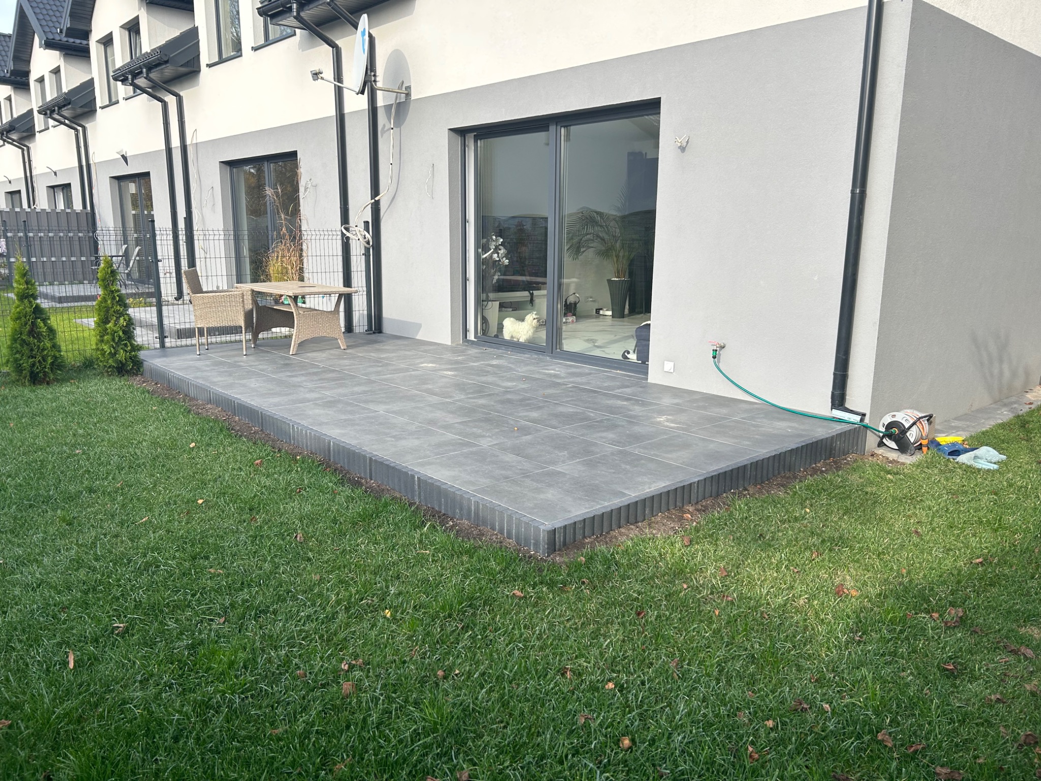 Nowoczesny taras z szarej kostki brukowej na trawniku, z rattanowymi meblami ogrodowymi i widokiem na dom z przesuwnymi drzwiami tarasowymi.