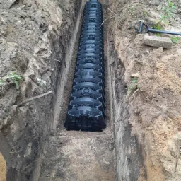 Długi rów w ziemi z zainstalowanymi modułami drenażowymi z tworzywa sztucznego, zasypanymi drobnym żwirem, szpadel i fragment rury PCV w tle.