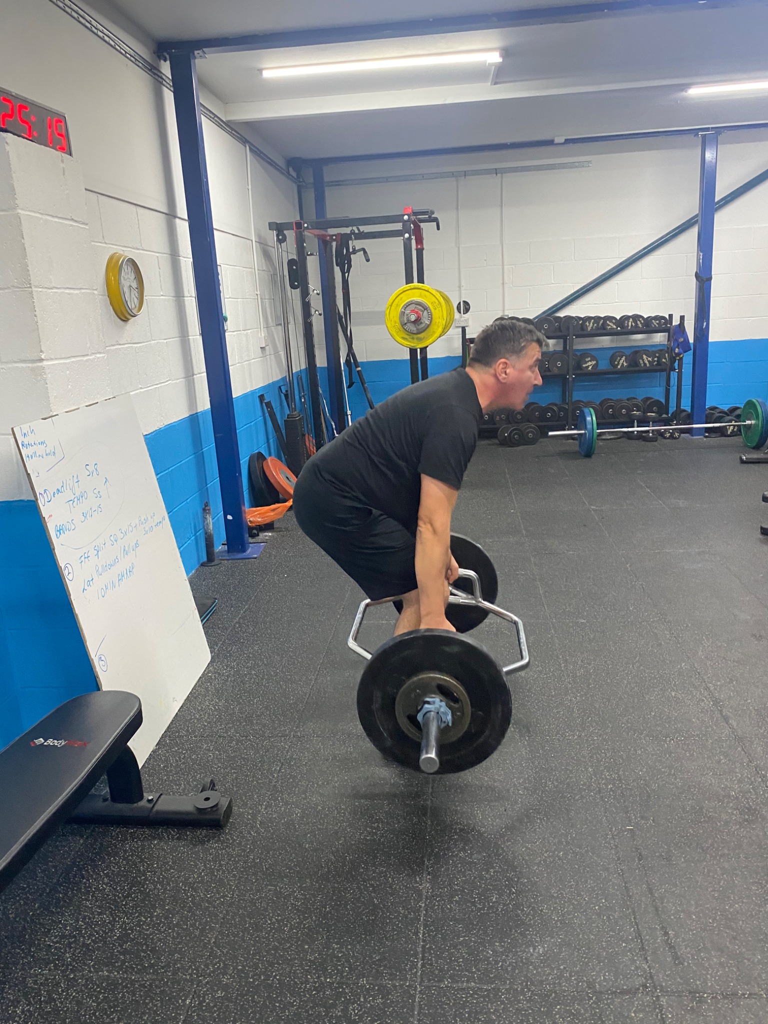 Mężczyzna w czarnym stroju wykonuje martwy ciąg na trap barze w siłowni; tablica z zapisanym planem treningowym w tle.