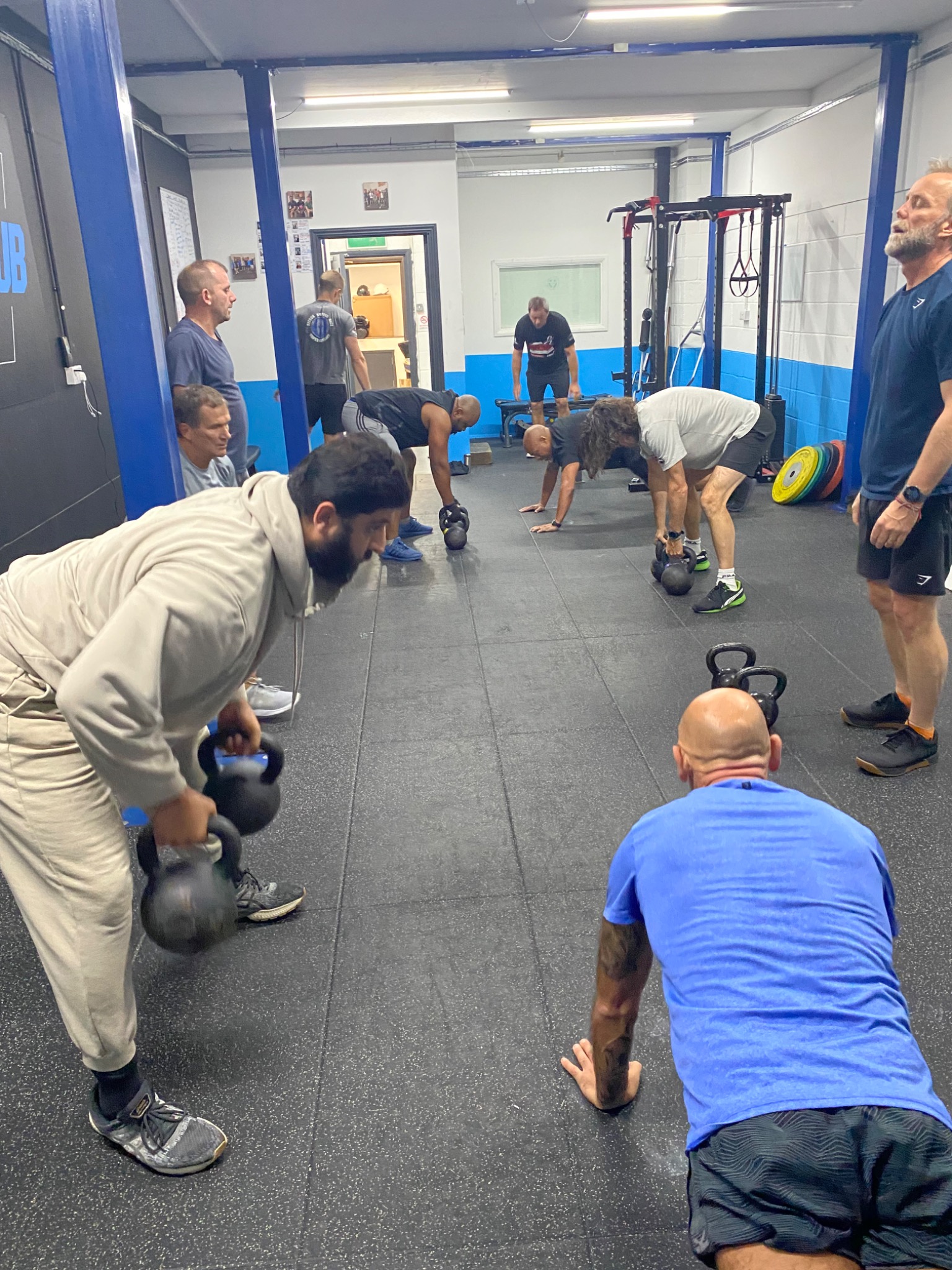 Grupa mężczyzn ćwiczy z odważnikami kettlebell w siłowni, pod okiem trenera, w tle widoczny sprzęt do ćwiczeń i osoby obserwujące.