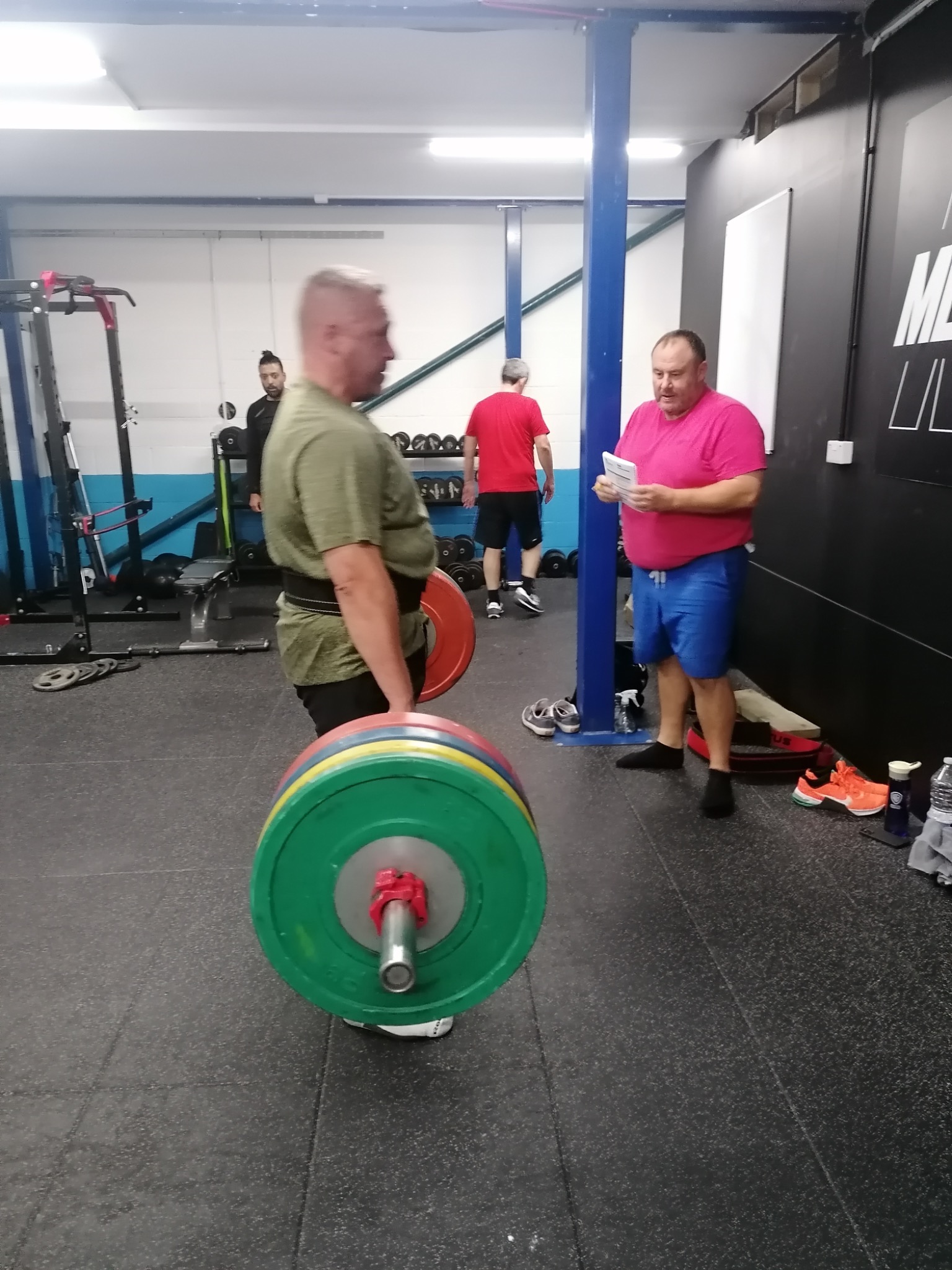 Mężczyzna w pasie kulturystycznym podnosi sztangę z kolorowymi obciążeniami w siłowni, drugi mężczyzna w różowej koszulce obserwuje z kartką w ręku.