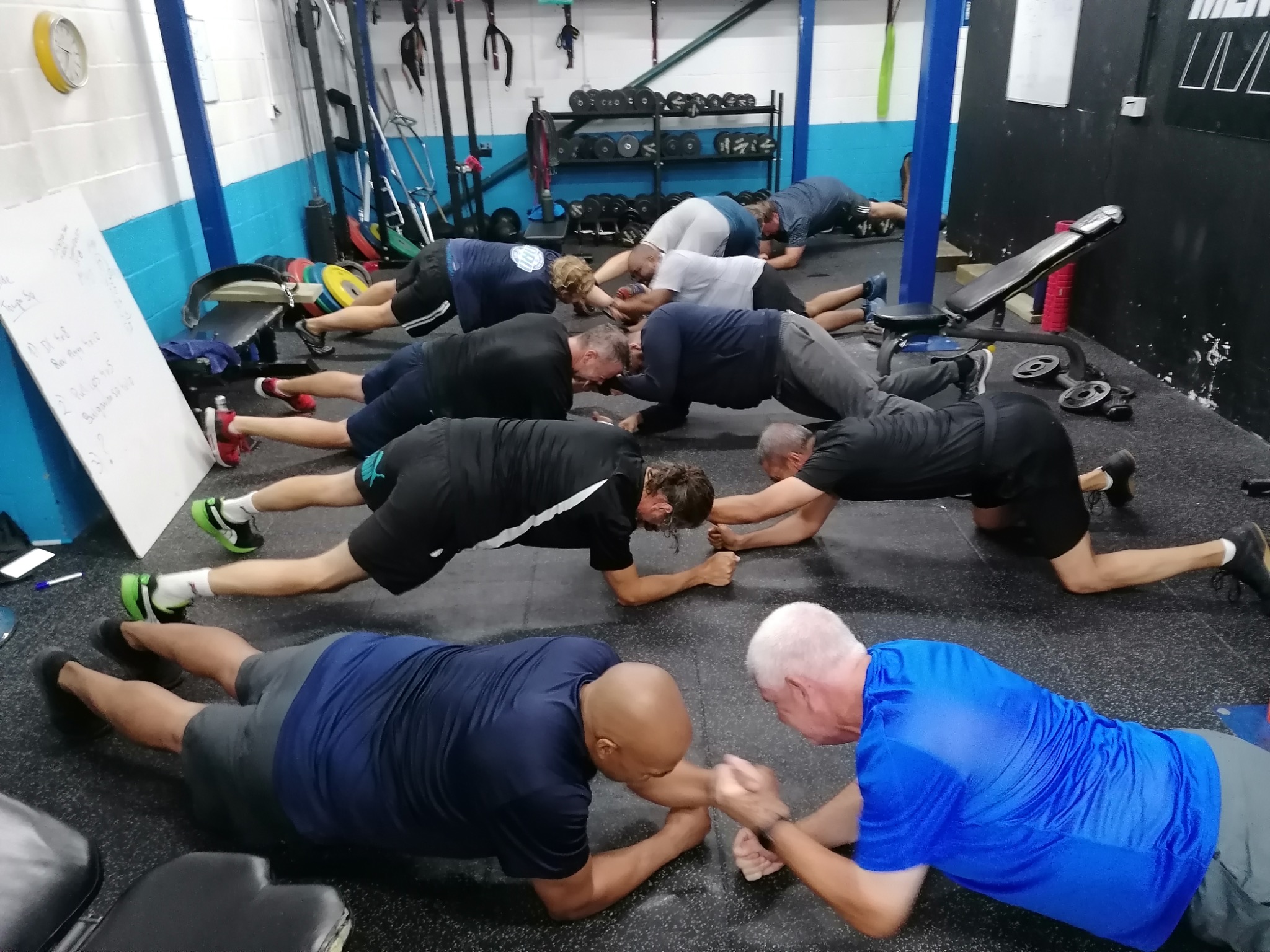 Grupa mężczyzn w różnym wieku wykonuje ćwiczenie plank w siłowni, na podłodze widoczne ślady użytkowania, w tle sprzęt do ćwiczeń i tablica z zapisanym planem treningowym.