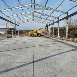 KOSTAL Konrad Kosikowski - Montaż stalowej konstrukcji hali z użyciem podnośnika teleskopowego. Widoczne spawanie elementów na wysokości, betonowa posadzka i błękitne niebo w tle.