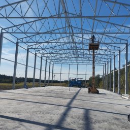 KOSTAL Konrad Kosikowski - Konstrukcja stalowa hali w trakcie montażu, widoczny podnośnik z pracownikiem na tle błękitnego nieba. Betonowa posadzka, widok z wnętrza hali.