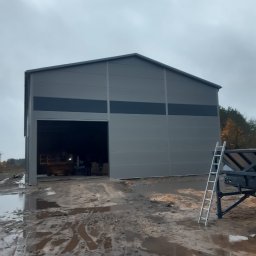 KOSTAL Konrad Kosikowski - Szara hala z segmentową bramą wjazdową, widoczna z zewnątrz na tle pochmurnego nieba. Na pierwszym planie błotniste podłoże i drabina oparta o przyczepę.