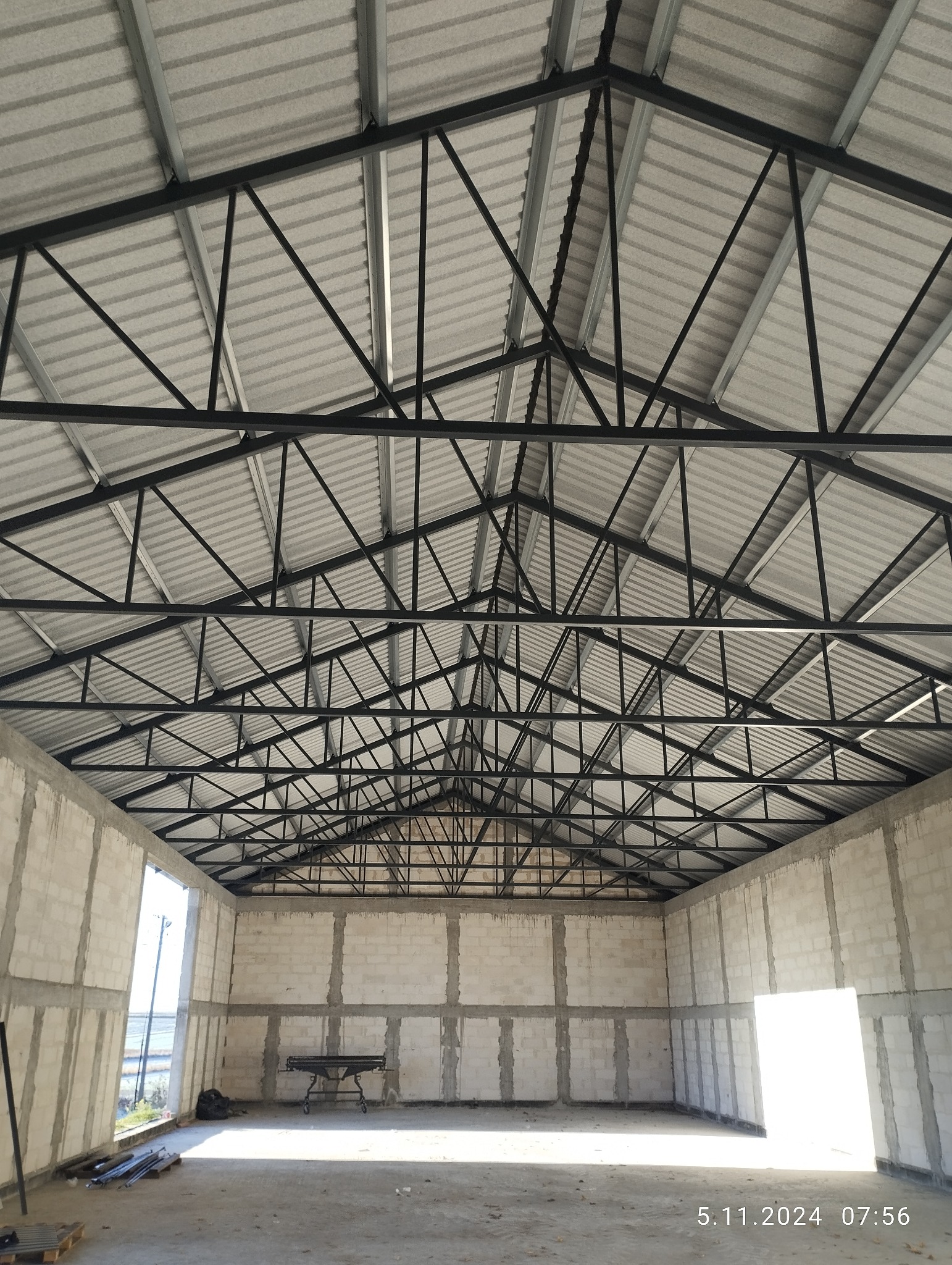 Wnętrze hali w budowie z widoczną konstrukcją dachu z metalowych belek i ścianami z betonu komórkowego. Widoczne narzędzia i elementy konstrukcyjne.