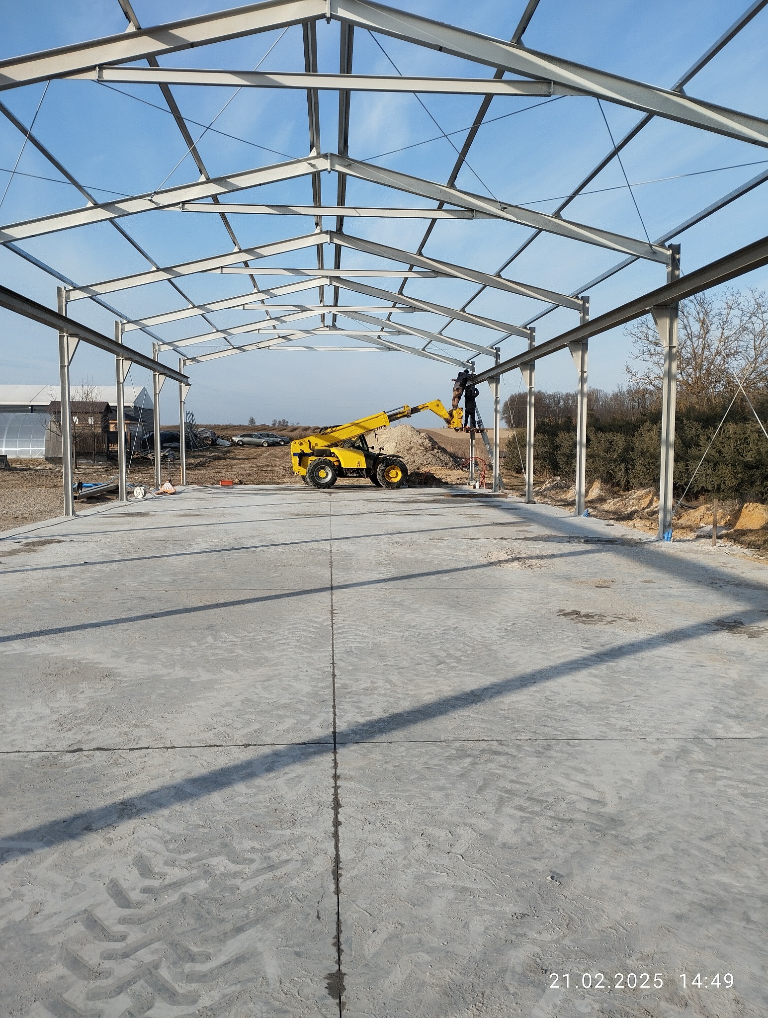 Montaż stalowej konstrukcji hali z użyciem podnośnika teleskopowego. Widoczne spawanie elementów na wysokości, betonowa posadzka i błękitne niebo w tle.