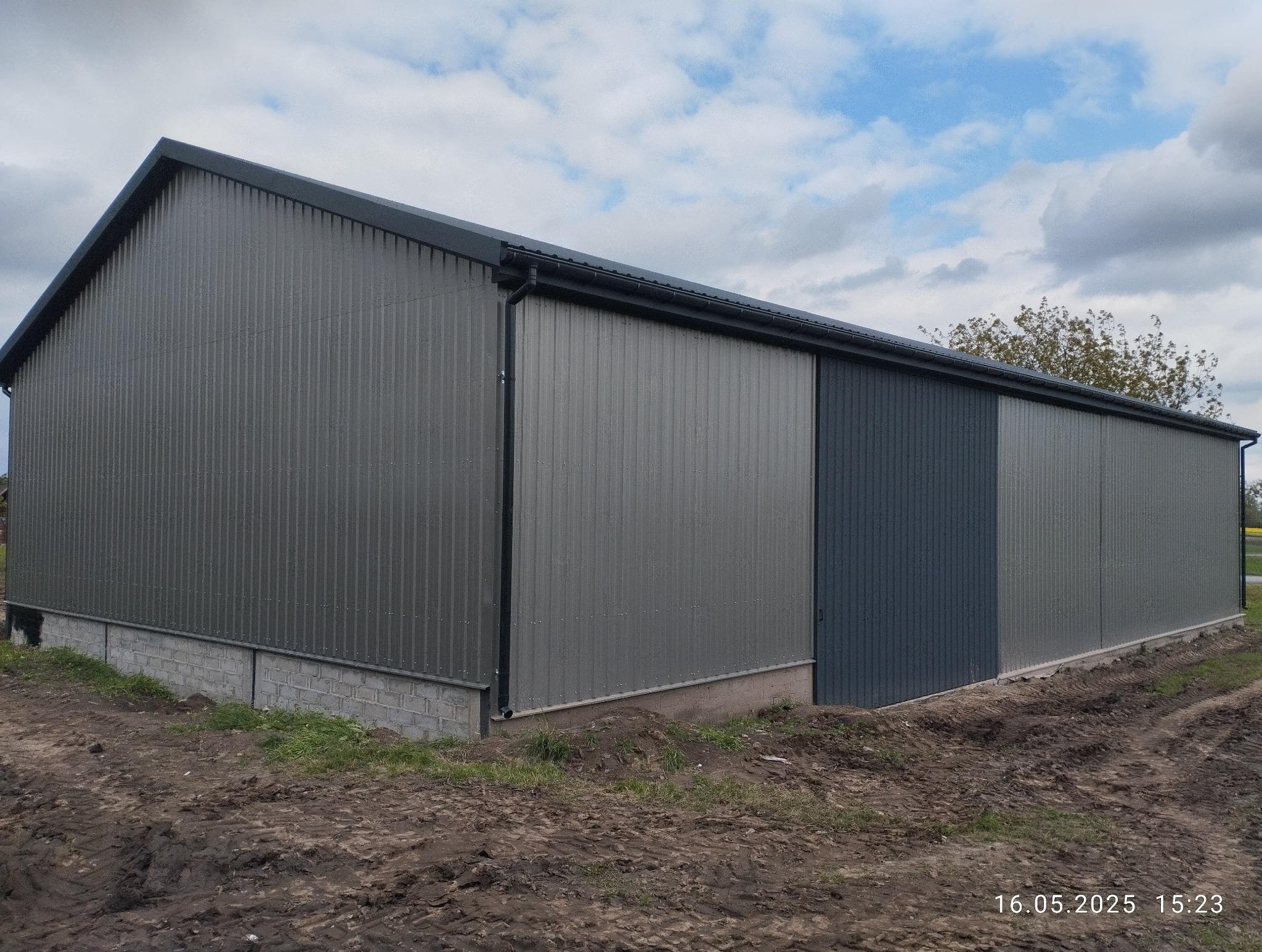 Szara hala z blachy trapezowej z widoczną bramą przesuwną, osadzona na fundamencie z bloczków betonowych. Tło: pochmurne niebo i fragment drzewa.