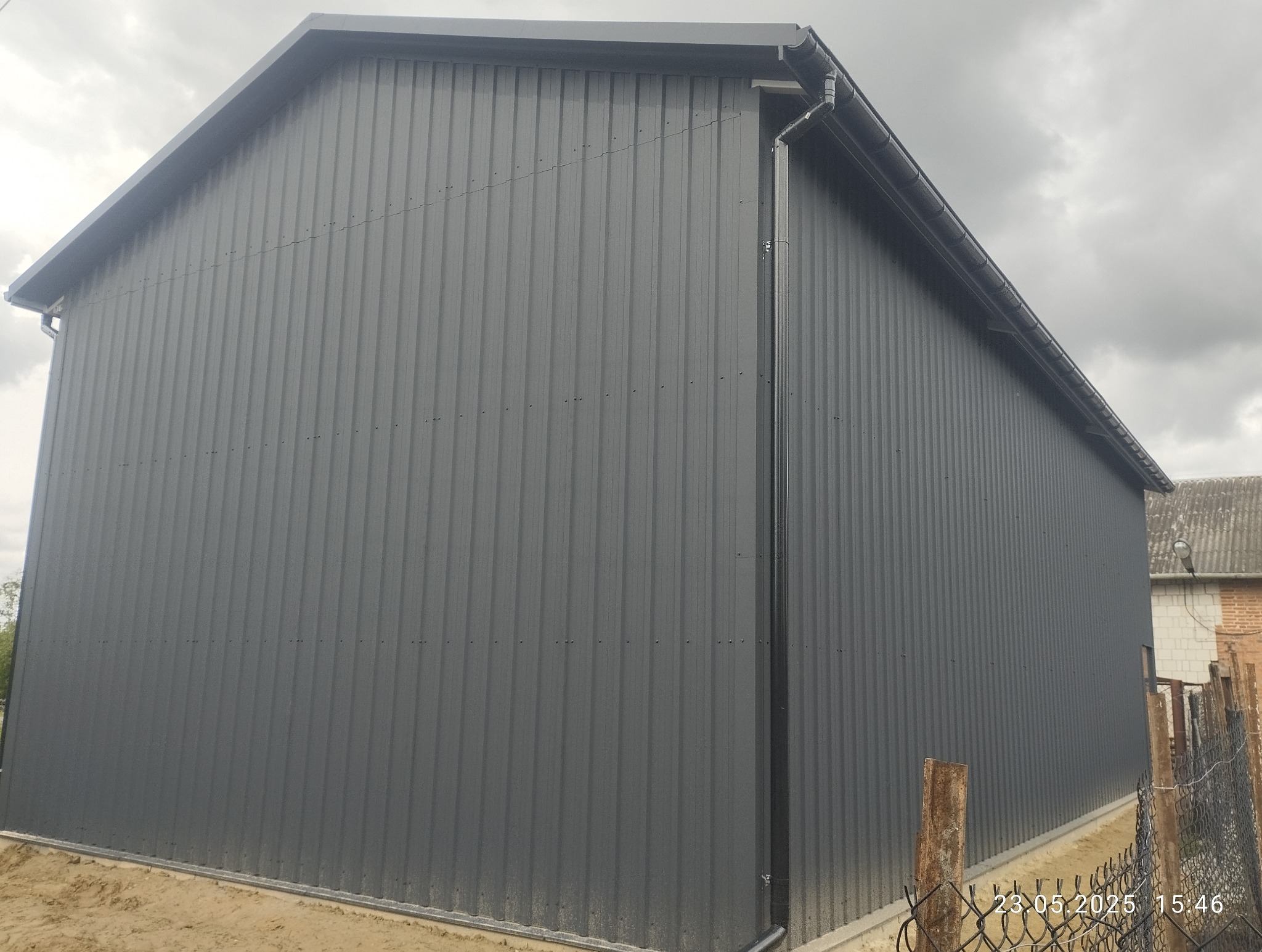 Nowoczesna hala z szarej blachy trapezowej, widok z zewnątrz. Detal: rynna spustowa wzdłuż ściany. Tło: pochmurne niebo i fragment sąsiedniego budynku.