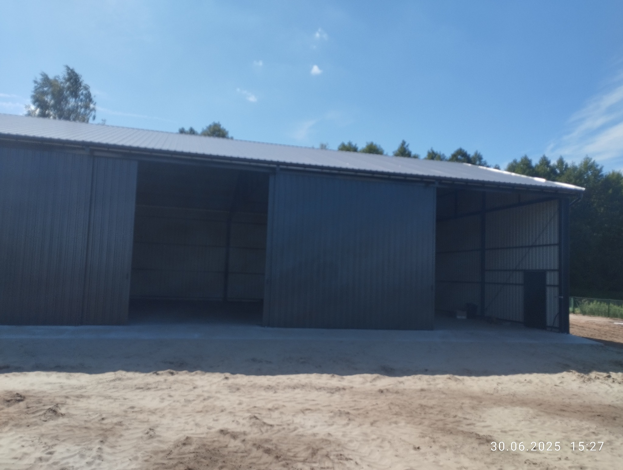 Nowoczesna hala stalowa z przesuwną bramą wjazdową, widok z zewnątrz w słoneczny dzień. Konstrukcja z szarej blachy trapezowej na betonowej wylewce.