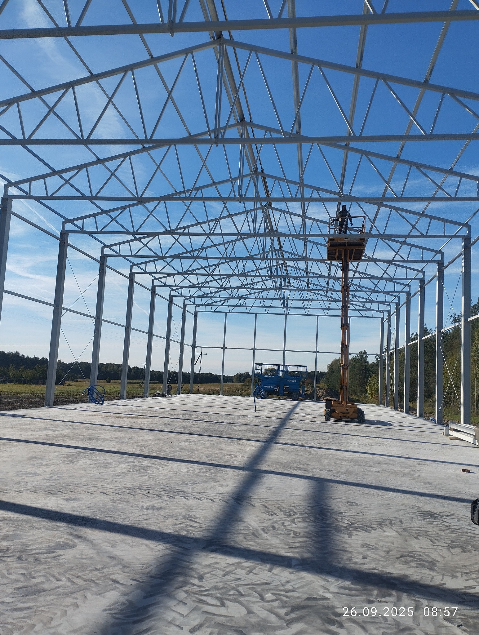 Konstrukcja stalowa hali w trakcie montażu, widoczny podnośnik z pracownikiem na tle błękitnego nieba. Betonowa posadzka, widok z wnętrza hali.