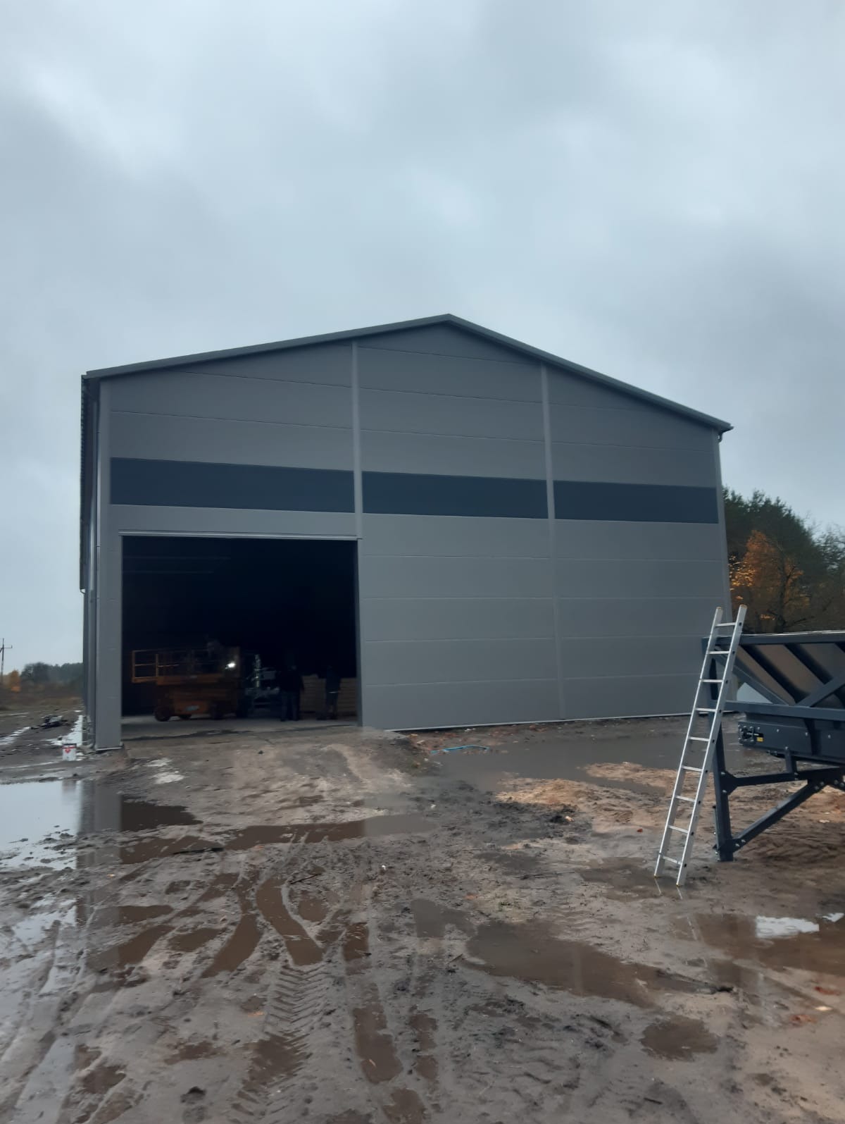 Szara hala z segmentową bramą wjazdową, widoczna z zewnątrz na tle pochmurnego nieba. Na pierwszym planie błotniste podłoże i drabina oparta o przyczepę.