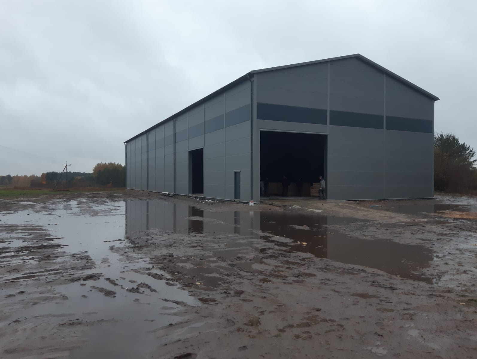 Szara hala stalowa z otwartą bramą wjazdową, widoczna w pochmurny dzień. Na pierwszym planie błotniste podłoże z kałużami, w tle drzewa z jesiennymi liśćmi.