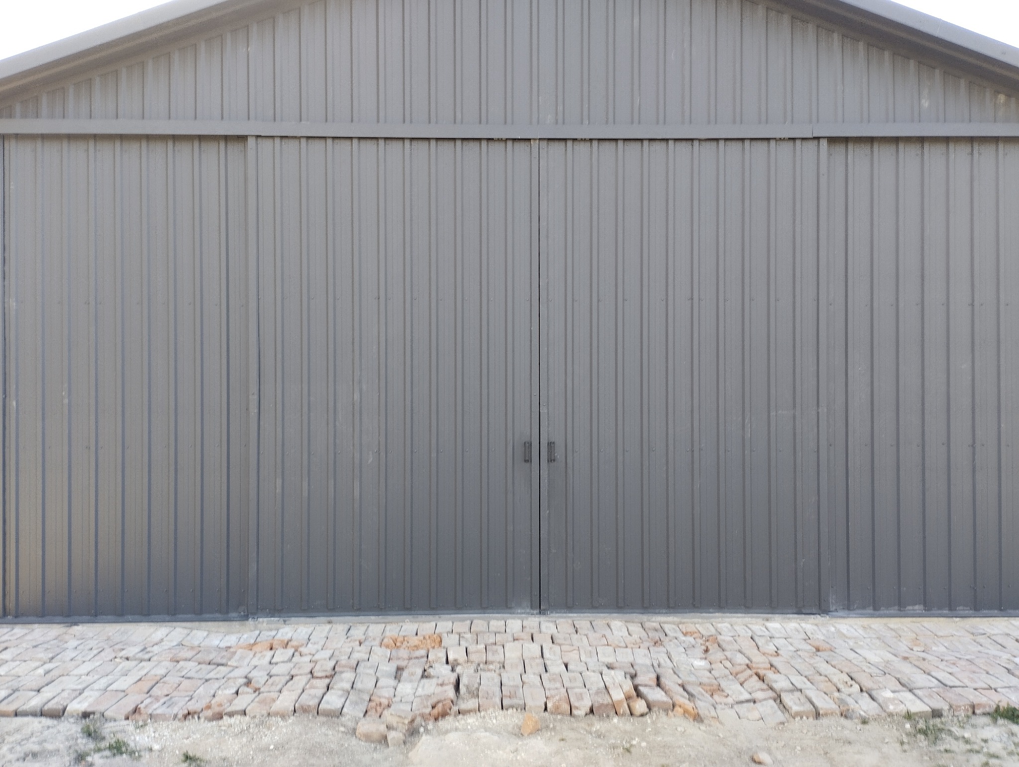 Szara, dwuskrzydłowa brama hali z blachy falistej, z widocznymi uchwytami, na tle ceglanej posadzki. Ujęcie centralne, symetryczne.