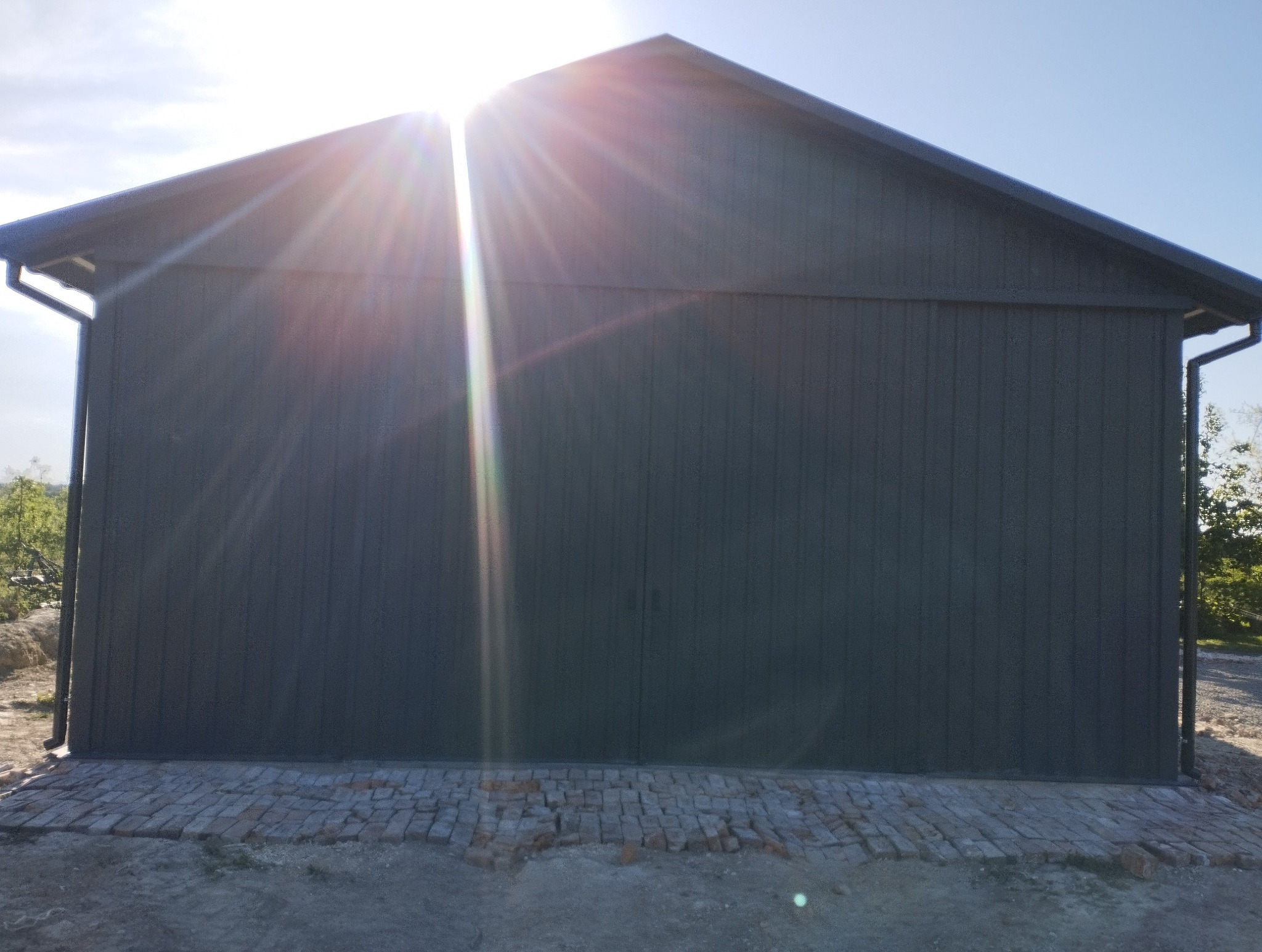 Nowoczesna, ciemna hala z blachy trapezowej, oświetlona promieniami słońca. Widoczny fragment fundamentu z cegły i orynnowanie. Minimalistyczny design.