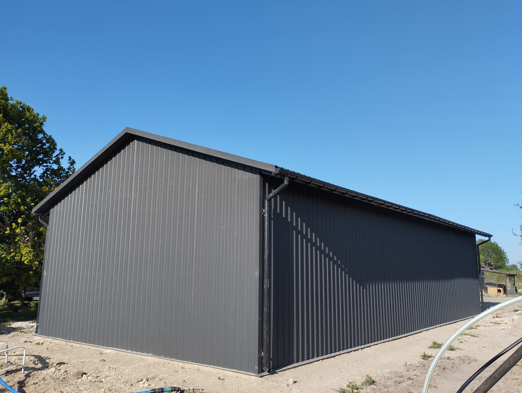 Szara hala z blachy trapezowej w słoneczny dzień. Widoczny fragment dachu z rynnami i drzewo w tle. Ujęcie z niskiej perspektywy podkreśla rozmiar konstrukcji.