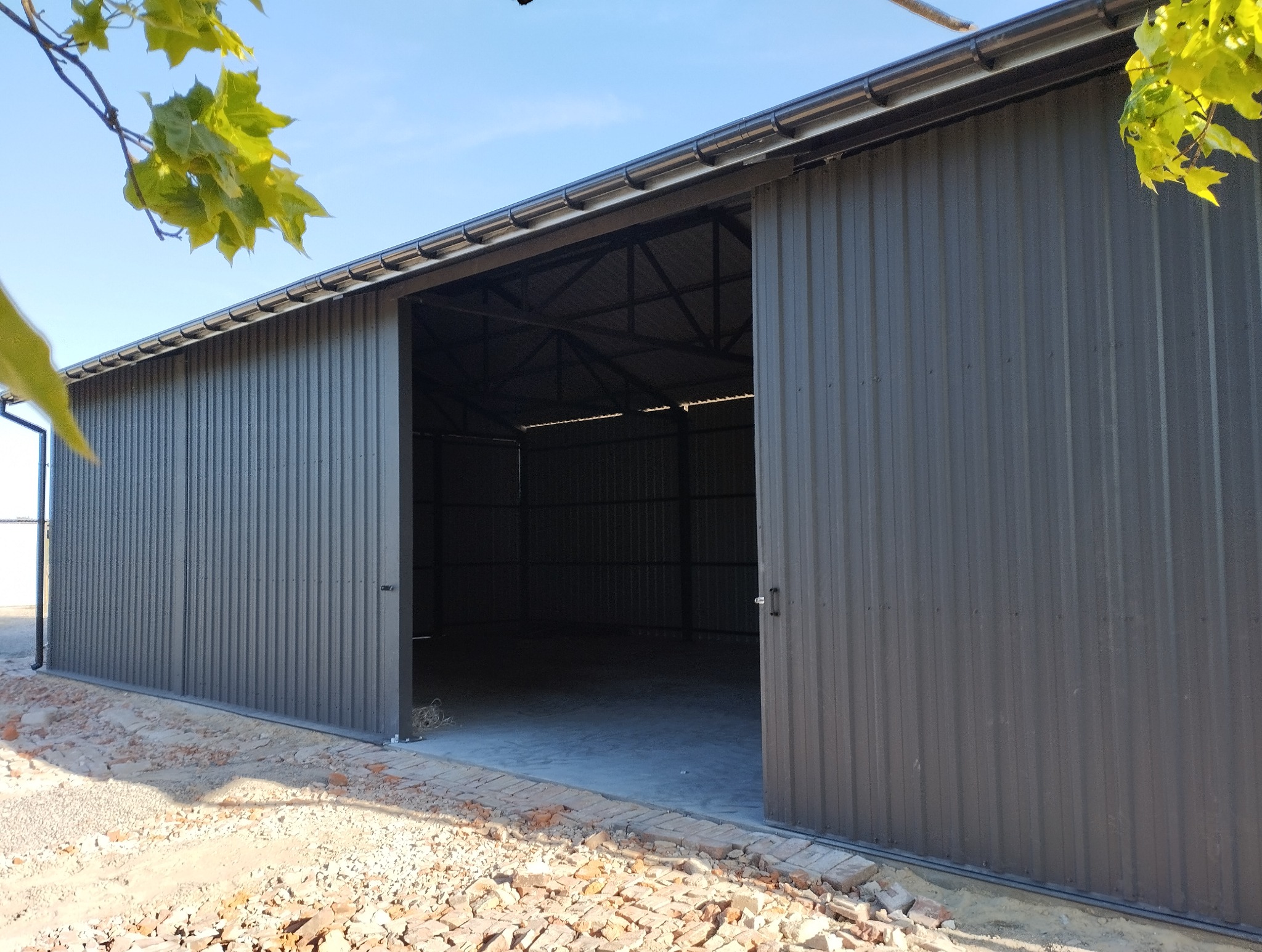 Otwarta, szara hala z blachy falistej w słoneczny dzień. Widoczne detale konstrukcji dachu i przesuwne drzwi. Fragmenty zielonych liści w górnych rogach kadru.