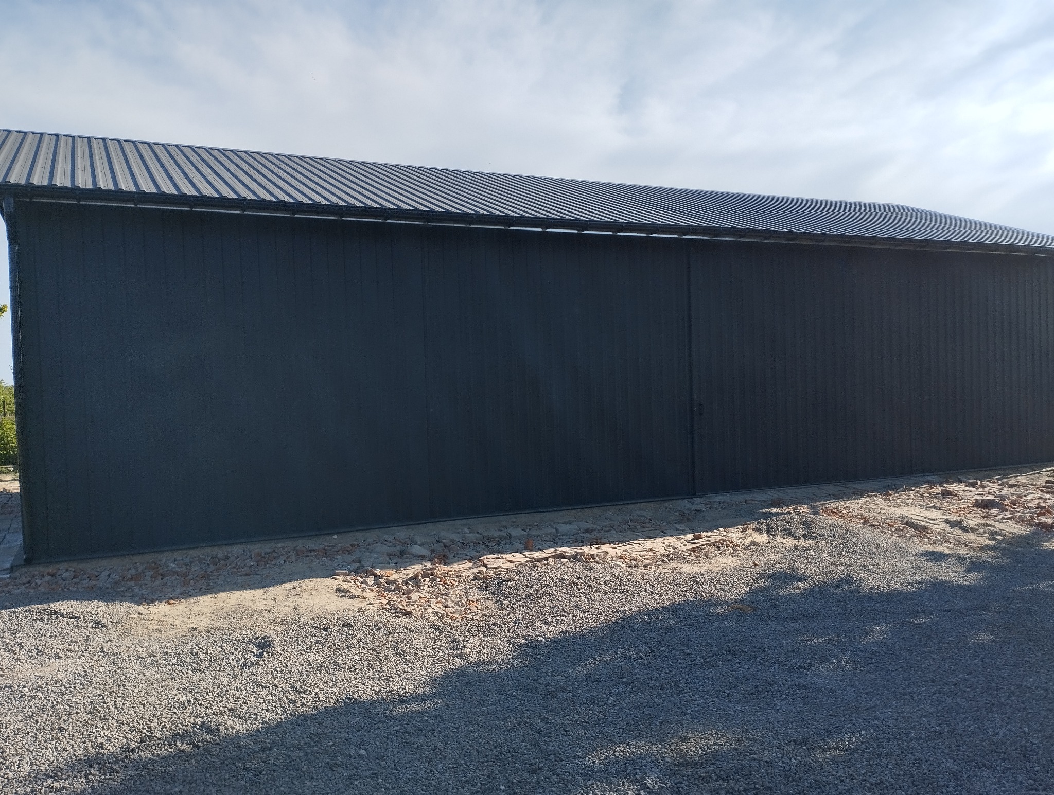 Szara hala z blachy trapezowej z widocznym dachem i fragmentem terenu z gruzem i kamieniami. Hala o dużej powierzchni, z widocznymi łączeniami blach.