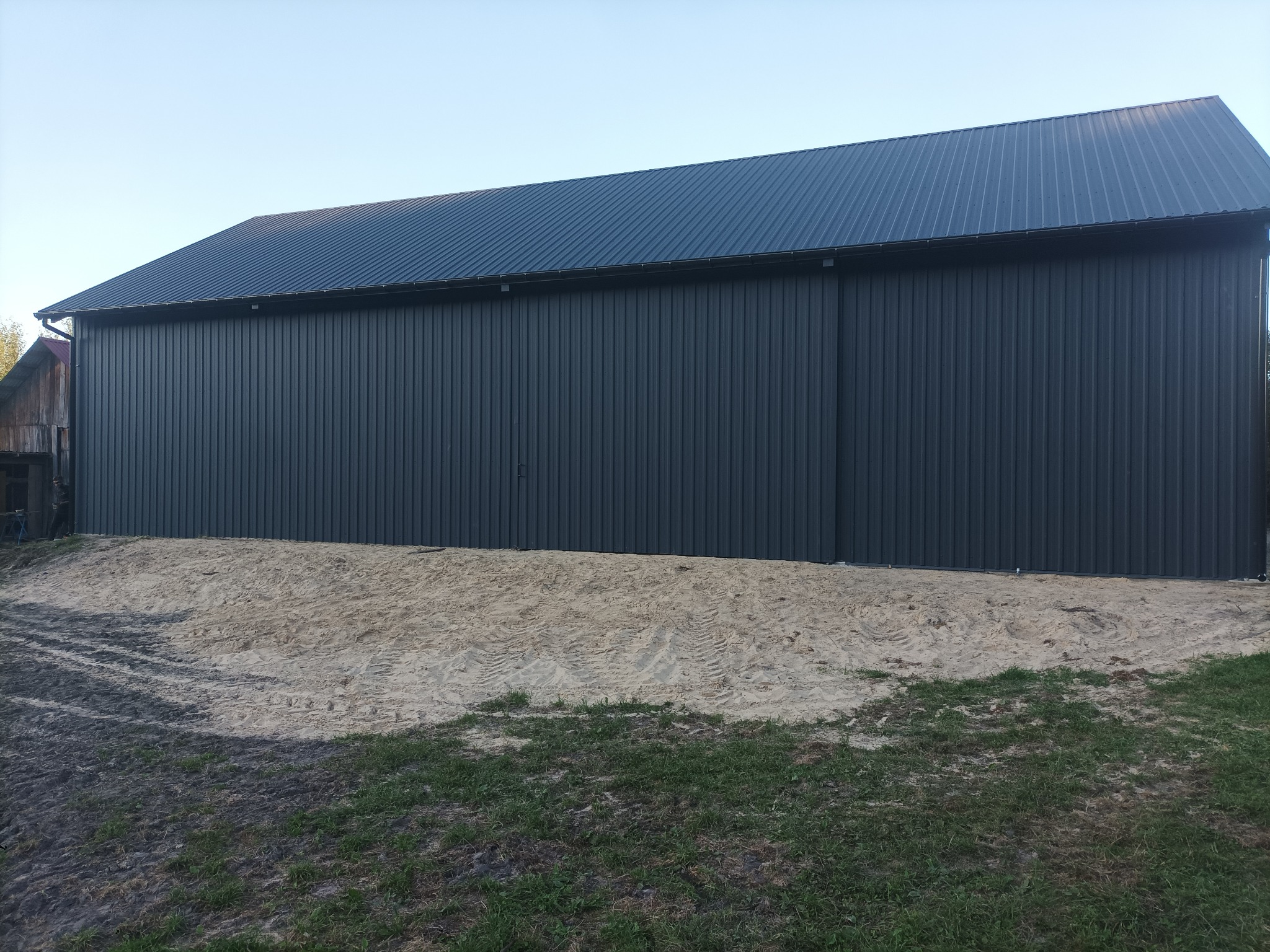 Nowoczesna hala stalowa z szarej blachy trapezowej, widok z zewnątrz. Ślady opon na piasku przed budynkiem. Częściowo widoczny budynek gospodarczy obok.