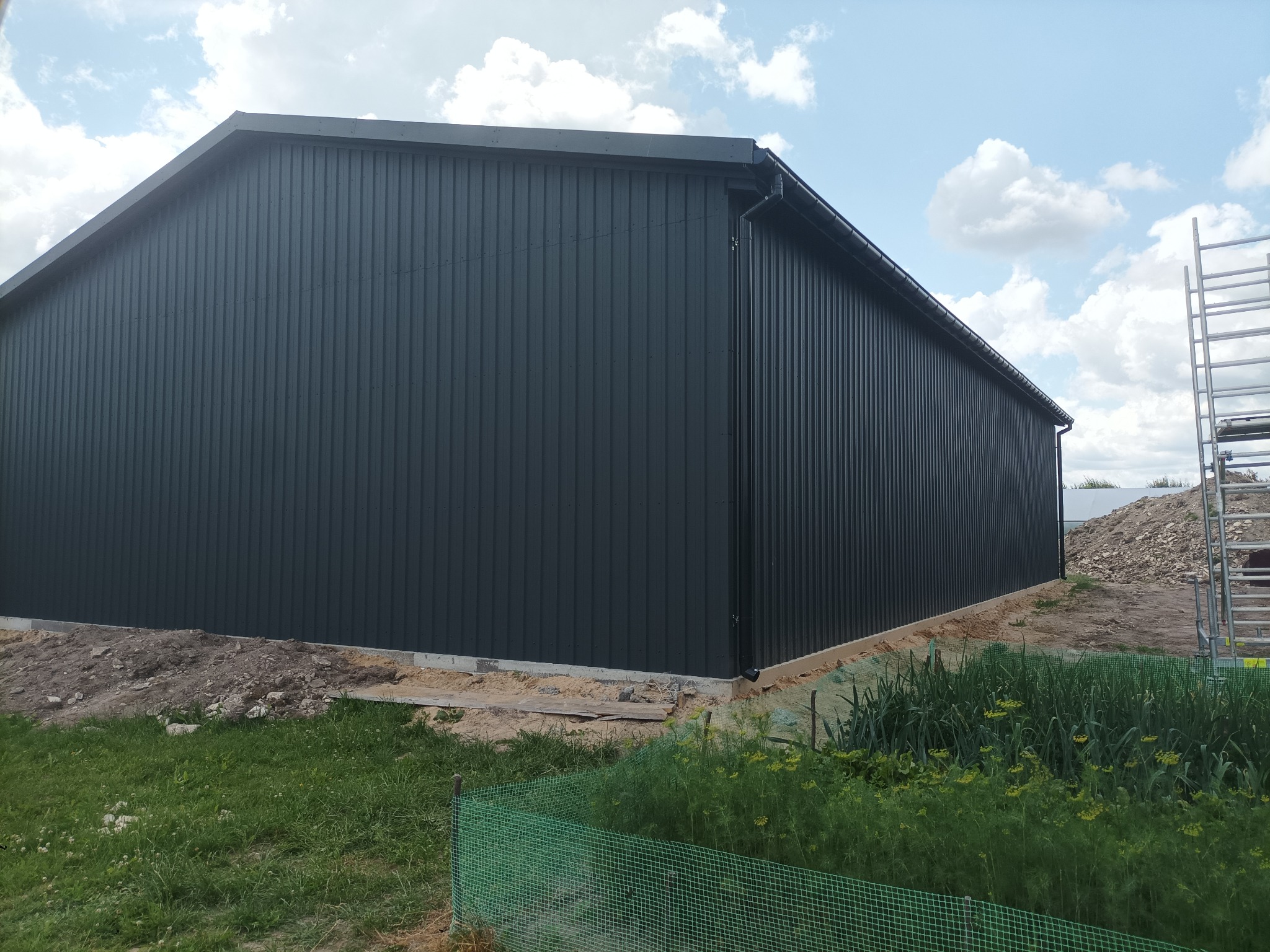 Nowoczesna hala z czarnej blachy trapezowej, widok z zewnątrz. Widoczne detale wykończenia, trawa i drabina oparta o ścianę. Błękitne niebo z chmurami w tle.