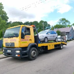 Żółta laweta MAN z napisem 'Pomoc Drogowa' przewozi srebrny samochód osobowy na tle zieleni i błękitnego nieba z ptakami na liniach energetycznych.